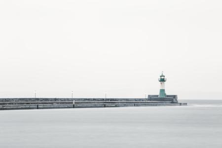 Das Bild zeigt einen ruhigen Küstenabschnitt mit einem langen Pier, an dessen Ende ein grün-weißer Leuchtturm steht. Mehrere Laternen säumen den Pier. Durch die helle, bewölkte Atmosphäre gehen Himmel und Meer nahezu nahtlos ineinander über. Die glatte Wasseroberfläche verstärkt die minimalistische und beruhigende Wirkung der Szenerie.