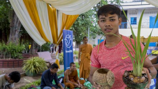 Ein junger Mann hält eine Pflanze und eine Kokusnuss in der Hand und schaut in die Kamera. Im Hintergrund sind buddistische Mönche und ein Banner von forumZFD. 